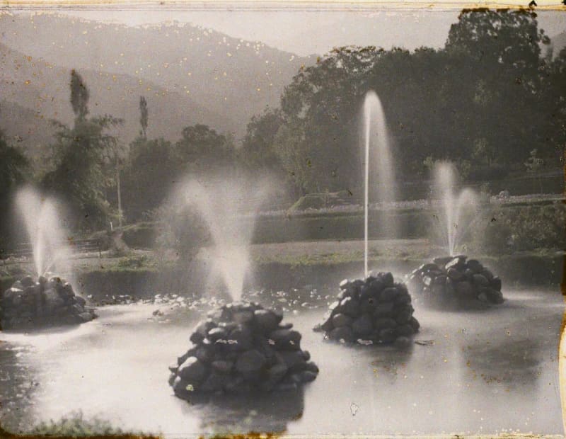 Fountains spraying from rock piles in a pond, backlit, with trees and hills behind.